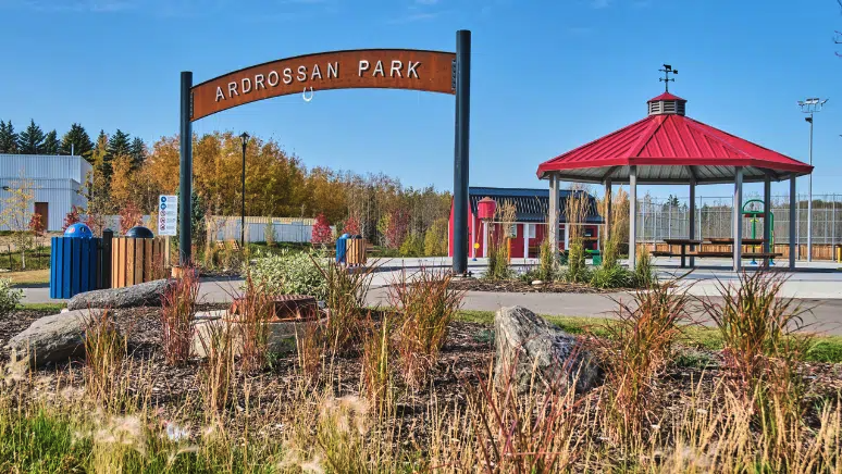 A scenic view of Ardrossan Park featuring a sign with the park's name, a red-roofed pavilion, and autumn-colored trees and plants under a clear blue sky.